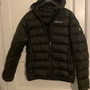 Svart dunjacka Peak Performance M - Svart dunjacka från Peak Performance med huva och broderad logga på bröstet. Jackan har quiltad design, dragkedja framtill och långa ärmar. Perfekt för kalla dagar och har en clean, sportig look.
