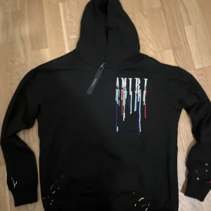 Svart hoodie från Amiri med färgstänk - Svart hoodie från Amiri med huva och coolt tryck på bröstet där loggan har färgstänk i vitt, blått, rött och gult som rinner ner. Tröjan har långa ärmar och detaljer med färgstänk även vid muddar och nederkant. Perfekt för dig som gillar streetwear och unik design.