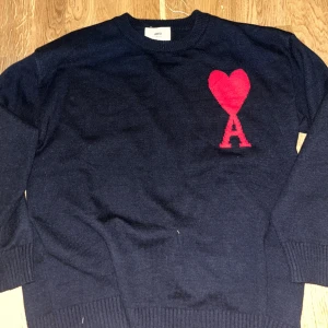 Mörkblå stickad tröja från Ami - Snygg mörkblå stickad tröja från Ami med rött hjärta och bokstaven A broderat på bröstet. Tröjan har rund hals, ribbade muddar och lång ärm. Perfekt för dig som gillar stilrena detaljer och vill ha något som sticker ut.