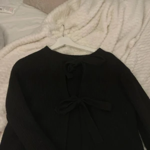 Svart stickad tröja med knyt - Svart stickad tröja med lång ärm och två snygga knytband framtill som ger en cool detalj. Tröjan har en enkel, rak passform och är perfekt för dig som gillar stilrena plagg med en twist. Mjuk och skön att bära under kyliga dagar.