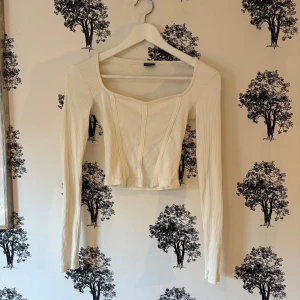 Vit croppad korsettopp Gina Tricot - Säljer en vit croppad korsettopp från Gina Tricot i ribbad bomull. Toppen har lång ärm, fyrkantig halsringning och markerade sömmar framtill som ger en snygg siluett. Perfekt att styla med höga jeans eller kjol för en trendig look.
