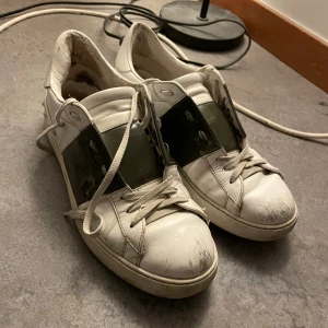 Valentino Garavani vita sneakers - Säljer ett par vita Valentino Garavani sneakers med bred olivgrön rem över snörningen. Skorna har rund tå, platt sula och är tillverkade i skinn. Snygg kontrast mellan vitt och olivgrönt, perfekt för dig som gillar exklusiva märken och streetstyle. Tyvärr har dom slitningar i skinnet på framsidan, sant ganska slitna inuti skon.