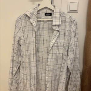 Vit rutig skjorta från Dressmann M - Vit skjorta från Dressmann i storlek M med tunna blå och svarta rutor. Klassisk krage, långärmad och bröstficka. Skjortan är i bomull och har knäppning framtill. Perfekt för en clean och stilren look.