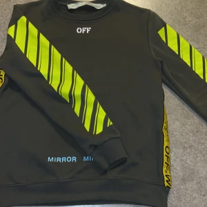 Off-White svart sweatshirt  - Svart sweatshirt från Off-White med neon-gula diagonala ränder och tryck på både fram- och baksida. Tröjan har rund hals, långa ärmar och coola detaljer som texten 'WHITE' och 'OFF' i vitt samt blått tryck nertill. Perfekt för dig som gillar streetwear och sticker ut.