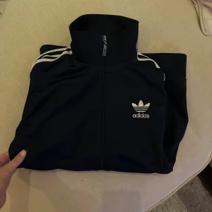 Adidas zipup  - Säljer en svart Adidas track jacket med klassiska vita ränder längs ärmarna och Adidas logga på bröstet. Jackan har hel dragkedja, hög krage och ribbade muddar. Lite slitage på metall detaljerna