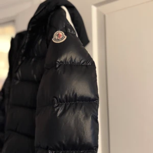 Svart Moncler pufferjacka med huva - Säljer en svart pufferjacka från Moncler med stor logga på huvan och klassisk Moncler-patch på ärmen. Jackan har dragkedja framtill, quiltad design och huva. Perfekt för dig som vill ha en snygg och varm jacka med streetkänsla.