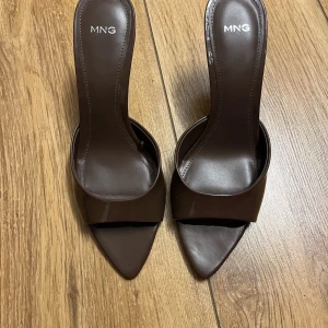 Klackar  - Snygga bruna klackar från MNG med öppen design och spetsig tå. Skorna har en hög klack och bred rem över foten. Tillverkade i glansigt syntetmaterial som ger en stilren look. Perfekta för dig som gillar minimalistisk och trendig stil.