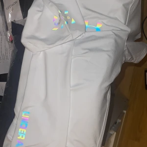 Vita Under Armour shorts med holografisk logga - Snygga vita shorts från Under Armour med en fet holografisk logga på benet. Perfekta för dig som gillar sportig stil och vill sticka ut lite extra. Sköna och lätta med elastisk midja, gjorda i ett syntetmaterial som andas.