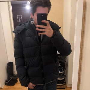 Superdry Pufferjacka mörkblå  - Säljer en mörkblå pufferjacka från Superdry med huva och dragkedja. Jackan har en clean look med diskret logga på ärmen och insidan. Perfekt för kalla dagar och riktigt skön passform. Fodrad och med praktiska fickor.