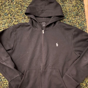 Svart hoodie från Polo Ralph Lauren - Svart hoodie från Polo Ralph Lauren med dragkedja framtill och klassisk huva. Tröjan har det ikoniska broderade logotypen på bröstet och praktiska fickor. Tillverkad i mjuk bomull, perfekt för en chill och stilren look.