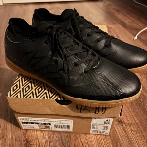 Umbro Velocita Alchemist 1.0 IC strl 42 - Snygga svarta inomhus-fotbollsskor från Umbro med gummisula och diskret mönster på ovandelen. Skorna har snörning och låg profil, perfekt för snabba rörelser på planen. Materialet är syntet och designen är stilren med svart ovandel och ljusbrun sula.