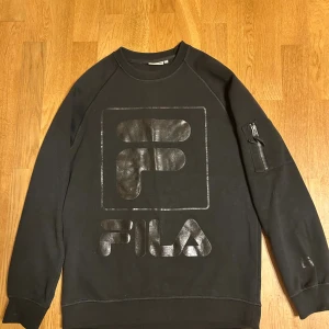 Svart sweatshirt FILA - Svart sweatshirt från FILA med stor glansig logga framtill. Tröjan har rund halsringning, lång ärm och en cool ficka med dragkedja på ena ärmen. Perfekt för dig som gillar streetwear och sportig stil.