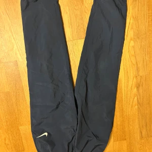 Blå Nike Dri-Fit träningsbyxor - Säljer ett par mörkblå Nike Dri-Fit träningsbyxor med elastisk midja och diskret Nike-logga på benet. Byxorna är tillverkade i lätt polyester och har meshfoder för extra ventilation. Perfekta för sport och aktivitet.