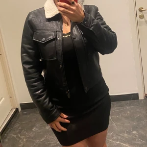 Svart skinnjacka med teddykrage - Säljer en svart skinnjacka med vit teddykrage och teddyfoder. Jackan har två stora fickor med lock och knappar framtill, samt dragkedja. Croppad modell med klassisk look, perfekt för dig som gillar streetstyle och vill ha en cool vibe.