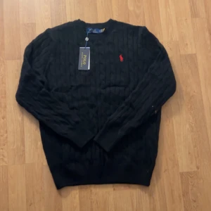 Svart kabelstickad tröja Polo Ralph Lauren - Svart kabelstickad tröja från Polo Ralph Lauren med röd broderad logga på bröstet. Rund hals och ribbade muddar vid ärmslut och nederkant. Klassisk och stilren design som funkar till allt. Perfekt för dig som gillar preppy vibes.