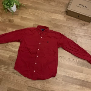Röd skjorta från Ralph Lauren - Snygg röd skjorta från Ralph Lauren med klassisk button-down krage och broderad logga på bröstet. Skjortan har långa ärmar, vita knappar och är gjord i mjuk bomull. Perfekt för dig som gillar stilrena plagg med ikonisk design.