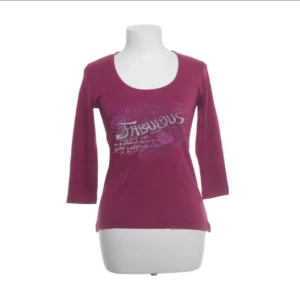 Lila y2k topp med tryck Fabulous - Snygg lila topp med trycket 'Fabulous' framtill och dekorativa detaljer i silver. Modellen har rund halsringning och trekvartsärm, perfekt för dig som gillar att sticka ut med färg och statement-tryck.