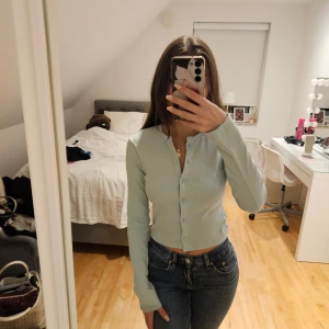 Mint grön  croppad kofta med knappar - Säljer en mint grön, croppad kofta med långa ärmar och knappar framtill. Tröjan har en ribbad struktur och sitter snyggt med en slim passform. Perfekt att styla med jeans eller kjol för en fräsch look.