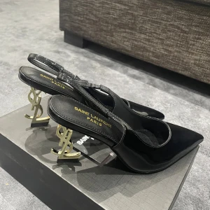 Saint Laurent svarta pumps med YSL-klack - Svarta pumps från Saint Laurent i blankt skinn med spetsig tå och öppen häl. Klacken är i guld och formad som YSL-loggan, vilket ger en lyxig och unik vibe. Snygg siluett och elegant design, perfekt för dig som gillar statement heels.