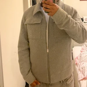 Grå overshirt med dragkedja - Snygg grå overshirt med rak passform och stora bröstfickor. Jackan har en rejäl dragkedja framtill, klassisk krage och långa ärmar med knapp vid ärmslut. Materialet är mjukt och känns som ull eller syntet, perfekt för lager-på-lager-stil.