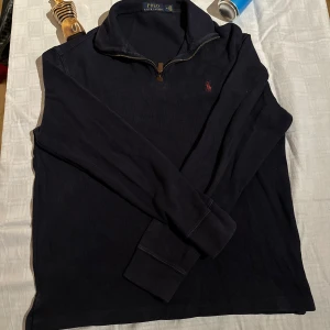 Marinblå half zip tröja Polo Ralph Lauren - Snygg marinblå långärmad tröja från Polo Ralph Lauren med klassisk röd logga på bröstet. Tröjan har en half zip-dragkedja vid kragen och är gjord i mjuk bomull. Perfekt för lager-på-lager och chill stil. Obs nocco och parfym följer ej med