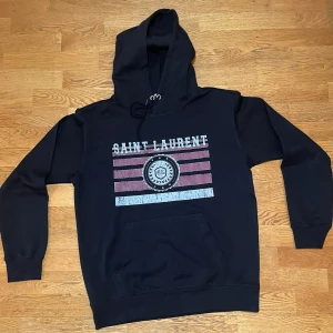 Svart YSL Hoodie - Svart hoodie från Saint Laurent Paris med tryck i vitt och rött framtill, använd fåtal gånger. Klassisk känguruficka, snörning i huvan och ribbade muddar. Perfekt för dig som gillar streetstyle och vill ha något snyggt och bekvämt. Skriv privat för mer information.