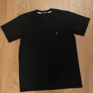 Svart t-shirt från Polo Ralph Lauren - Svart t-shirt från Polo Ralph Lauren med klassisk röd logobrodering på bröstet. Modellen har rund hals och korta ärmar, perfekt för dig som gillar stilrena och enkla plagg med en touch av lyx.