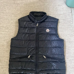 Svart dunväst från Moncler - Svart dunväst från Moncler i storlek 1, med glansig finish och klassisk quiltad design. Västen har hög krage, två dragkedjefickor framtill och Moncler-logga på bröstet. Stängs med dragkedja hela vägen upp. Perfekt för lager-på-lager under kyliga dagar.