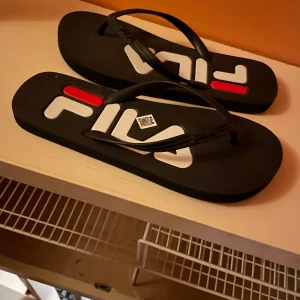 Svarta Fila sandaler med logga - Säljer ett par svarta sandaler från Fila med stor vit och röd logga på sulan. Klassisk flipflop-modell med svarta remmar och platt sula, perfekt för sommaren. Snygg och enkel design som passar till shorts eller badkläder.