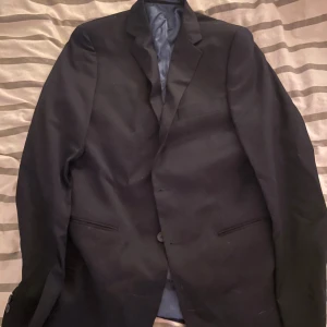 Svart kostym Dressmann slim fit - Snygg svart kostym från Dressmann i slim fit-modell. Kavajen har klassisk krage, två framfickor och knäppning med två knappar. Byxorna är raka och stilrena. Perfekt för dig som vill ha en enkel och stilfull look. (ANVÄND EN GÅNG)!
