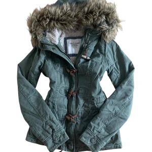 Grön vintage parkajacka Abercrombie & Fitch - Snygg grön parkajacka från Abercrombie & Fitch med fuskpäls på huvan och mysigt teddyfoder. Jackan har långa ärmar, snörning i midjan och coola träknappar framtill. Perfekt för dig som gillar streetstyle och vill hålla dig varm under hösten och vintern. Vintage from the 2000s/2010s!! 