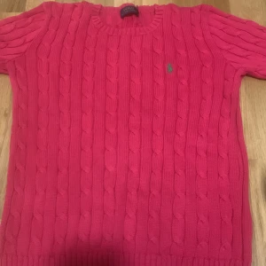 Rosa stickad tröja från Polo Ralph Lauren - Säljer en snygg rosa stickad tröja från Polo Ralph Lauren med klassiskt kabelstickat mönster och rund hals. Tröjan har korta ärmar och den ikoniska gröna Polo-loggan broderad på bröstet. Perfekt för dig som gillar färg och stil.