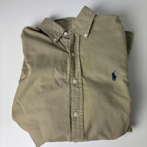Beige skjorta från Ralph Lauren XS - Stilren beige skjorta från Ralph Lauren i slim fit-modell. Skjortan har klassisk krage, vita knappar och lång ärm. På bröstet finns den ikoniska blå broderade loggan. Materialet är mjuk bomull som känns skönt mot huden.