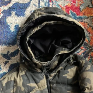 Camouflage pufferjacka från Zara - Säljer en pufferjacka från Zara med camouflage-mönster i gröna och beige toner. Jackan har huva, hel dragkedja framtill och är fodrad med svart polyester. Perfekt för kalla dagar och har en loose passform som ger skön streetkänsla.