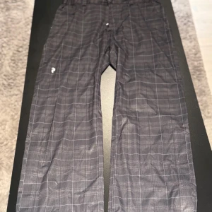 Rutmönstrade täckbyxor Peak Performance - Svarta täckbyxor från Peak Performance med diskret rutmönster i grått. Byxorna har rak passform och är tillverkade i ett slitstarkt syntetmaterial, perfekta för vinterns aktiviteter. Klassisk logga på benet och bekväm midja.