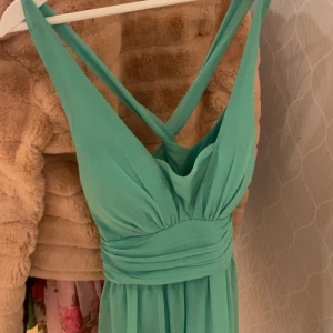 Turkos långklänning från NLY Eve - Säljer en turkos långklänning från NLY Eve med omlott-design och breda axelband som korsas i ryggen. Klänningen har ett snyggt fall och markerad midja med draperade detaljer. Materialet är lätt och känns luftigt, perfekt för varmare dagar.