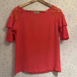 Röd rosa blus med spets och volangärmar - Säljer en snygg röd rosa  blus med spetsdetaljer på axlarna och dubbla volangärmar. Blusen har rund halsringning och är tillverkad i mjukt bomullsmaterial. Perfekt för dig som gillar färg och coola detaljer!