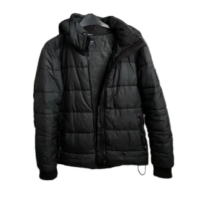 Svart pufferjacka Everest - Svart pufferjacka från Everest med huva och dragkedja framtill. Jackan har quiltad design och ribbade muddar vid ärmslut. Perfekt för kalla dagar och har en stilren look som funkar till det mesta.