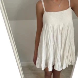 Sommarklänning Zara - Superfin vit kortklänning med smock upptill och tunna axelband. Klänningen har flera volanglager och en luftig, flowy passform som är perfekt för varma dagar. Tillverkad i ett lätt tyg som känns skönt mot huden.