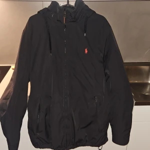Svart vindjacka från Polo Ralph Lauren - Svart vindjacka som är från Polo Ralph Lauren och är fodrad på insidan av jackan, röd broderad logga på bröstet. Jackan har huva med snörning, dragkedja framtill och två sidofickor med dragkedja en ficka på insidan. Perfekt för dig som gillar stilren och sportig look.