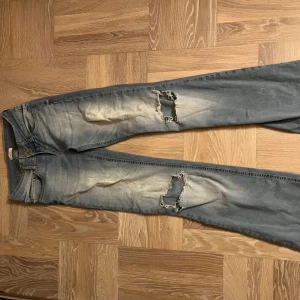 Bootcut jeans med slitningar från Nelly - Bootcut jeans från Nelly i ljusblå tvätt med slitna detaljer och hål på båda benen. Klassisk femficksmodell med knappstängning och snygga bakfickor med lock. Jeansen har en cool, lite vintage vibe och är gjorda i mjukt denimtyg.