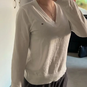 Vit v-ringad tröja Tommy Hilfiger - Vit långärmad tröja från Tommy Hilfiger med v-ringning och liten broderad logga på bröstet. Tröjan är i mjuk bomull och har en klassisk, clean look som passar perfekt till jeans eller kjol.