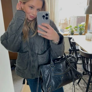 Grå quiltad jacka från Zara - Quiltad jacka i mörkgrått från Zara med loose passform, två stora fickor framtill och dragkedja. Jackan har ett vågigt mönster i quiltningen och står krage. Perfekt för dig som gillar chill och trendig stil. Storlek S