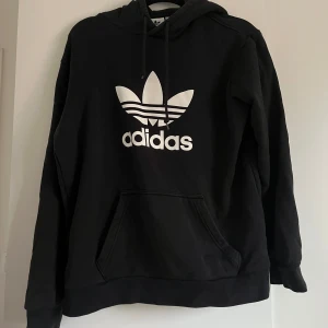 Svart Adidas hoodie - Svart hoodie från Adidas. Klassisk känguruficka framtill, dragsko i huvan och ribbade muddar. 