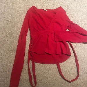 Röd omlottblus med knytband - Säljer en snygg röd omlottblus med långa ärmar och knytband i midjan. Blusen har v-ringad hals och ett lätt ribbat tyg som ger en cool struktur. Perfekt för dig som gillar att sticka ut med färg och vill ha en trendig topp med lite extra detaljer.