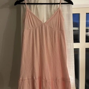 Ljusrosa kortklänning med volang - Supersöt ljusrosa klänning med tunna axelband och v-ringad front. Klänningen har en flowy passform, volangdetaljer och smockad rygg för extra komfort. Materialet är lätt och krinklat, perfekt för varma dagar. Söt spetsdekor längs volangerna ger en extra touch.