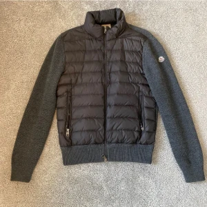 Moncler grå Cardigan - Snygg pufferjacka från Moncler i mörkgrå färg med quiltad front och stickade ärmar. Jackan har ribbade muddar, två dragkedjefickor och Moncler-logga på vänster ärm. Perfekt för dig som gillar stilren design och vill ha något som sticker ut.