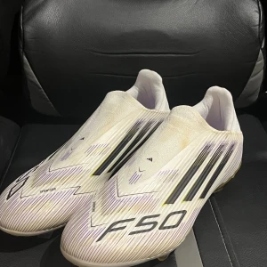 Adidas F50 fotbollsskor vit/gul - Säljer ett par Adidas F50 fotbollsskor i vitt med svarta och lila detaljer samt gul sula. Skorna har en strömlinjeformad design utan snörning, med mönstrade linjer och tre klassiska Adidas-ränder på sidan. Sulan har dobbar i gult och lila för grymt grepp på planen.