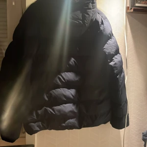 Svart pufferjacka från GANT - Säljer en svart pufferjacka från GANT med huva och dragkedja framtill. Jackan har ett stilrent, quiltat mönster och GANT-logga på ärmen. Perfekt för kalla dagar och har en schysst, modern look som funkar till streetwear nästan nyskick köptes för 1900