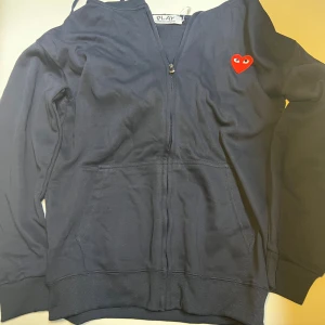 Mörkblå hoodie Comme des Garçons Play - Mörkblå hoodie från Comme des Garçons Play med dragkedja, huva och känguruficka. Ikoniskt rött hjärta med ögon broderat på bröstet. Skön passform och tillverkad i mjuk bomull. Perfekt för dig som gillar streetstyle och vill sticka ut.priset kan diskuteras vid snabbaffär.